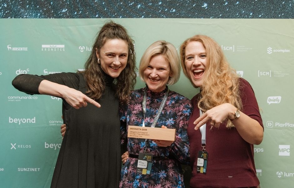 Drei Frauen vor einer Logowand zeigen auf ihren Preis